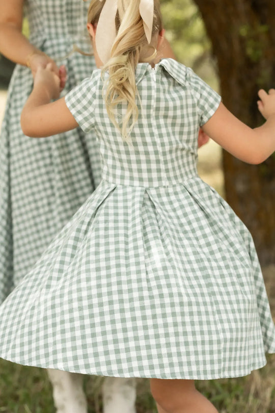 A young girl in the Ivy City Co Mini Amber Dress in Sage Gingham spins outdoors, holding hands with an adult wearing a matching cotton midi dress.