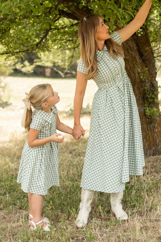 A woman and young girl wear matching Ivy City Co Mini Amber Dresses in Sage Gingham as they stand hand in hand under a tree on a grassy lawn, with the woman reaching up toward the branches.