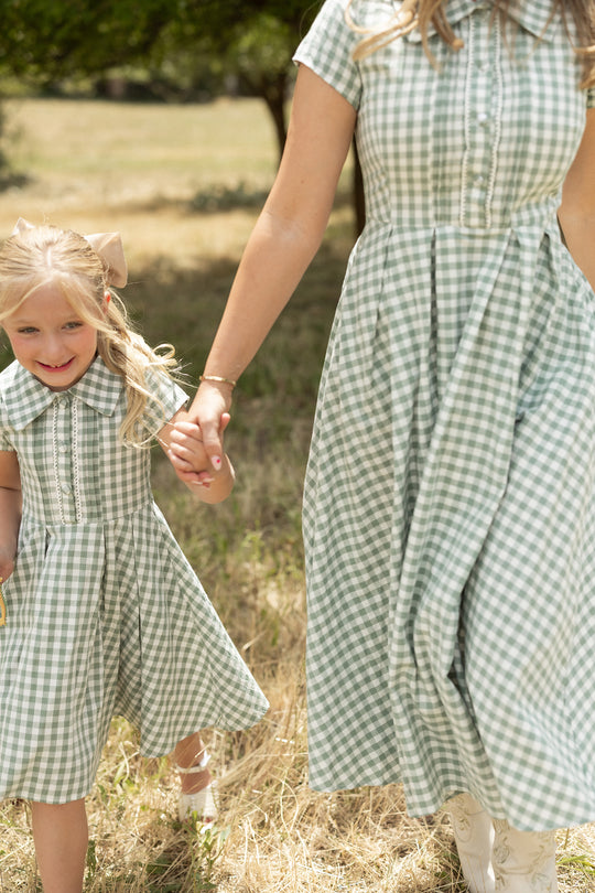 A woman and a young girl hold hands outdoors in a grassy field, both wearing Ivy City Co’s Mini Amber Dress in Sage Gingham with pleated skirts. The girl smiles as they walk together among the trees.