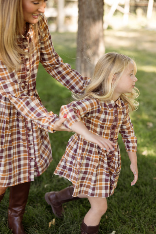 A woman and a young girl are outside on green grass, both wearing matching Ivy City Co Mini Bowie Long Sleeve Dresses in Spiced Plaid with brown boots. The woman gently holds the girl’s arm as she looks away, both styled in these charming cotton balloon sleeve dresses.