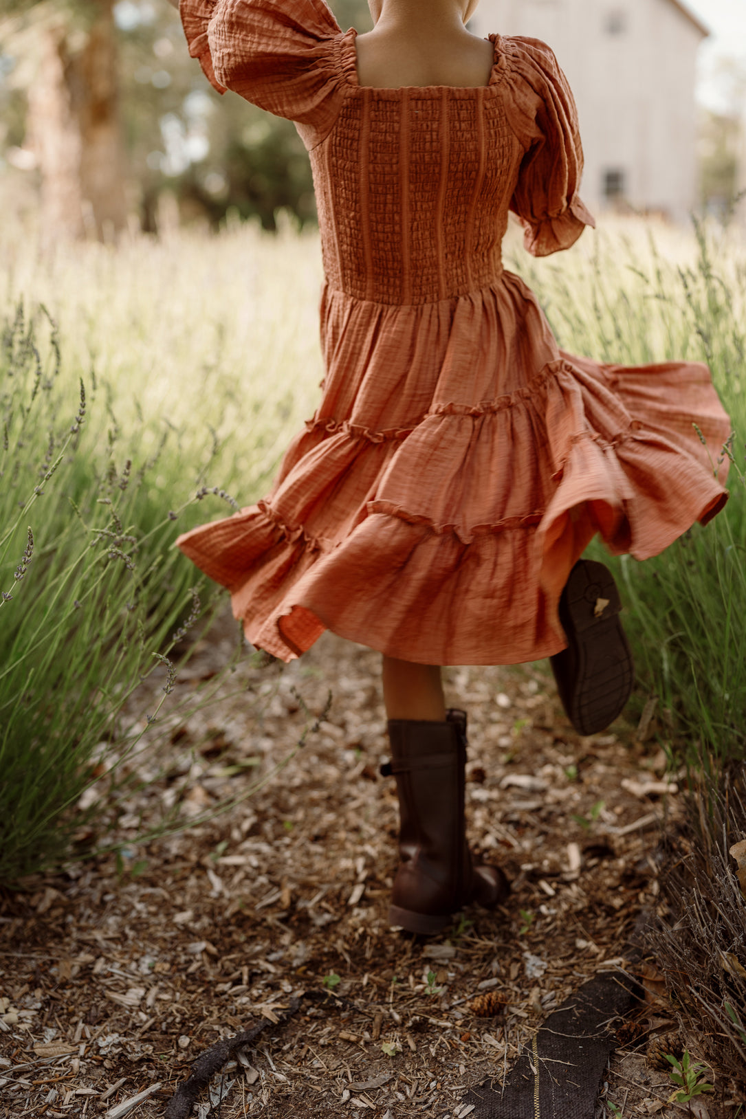 A child wearing the Ivy City Co Mini Madeline Dress in Apple Cider, featuring a smocked bodice and charming puff sleeves, twirls joyfully along a path bordered by tall grass, with a softly blurred building in the background.