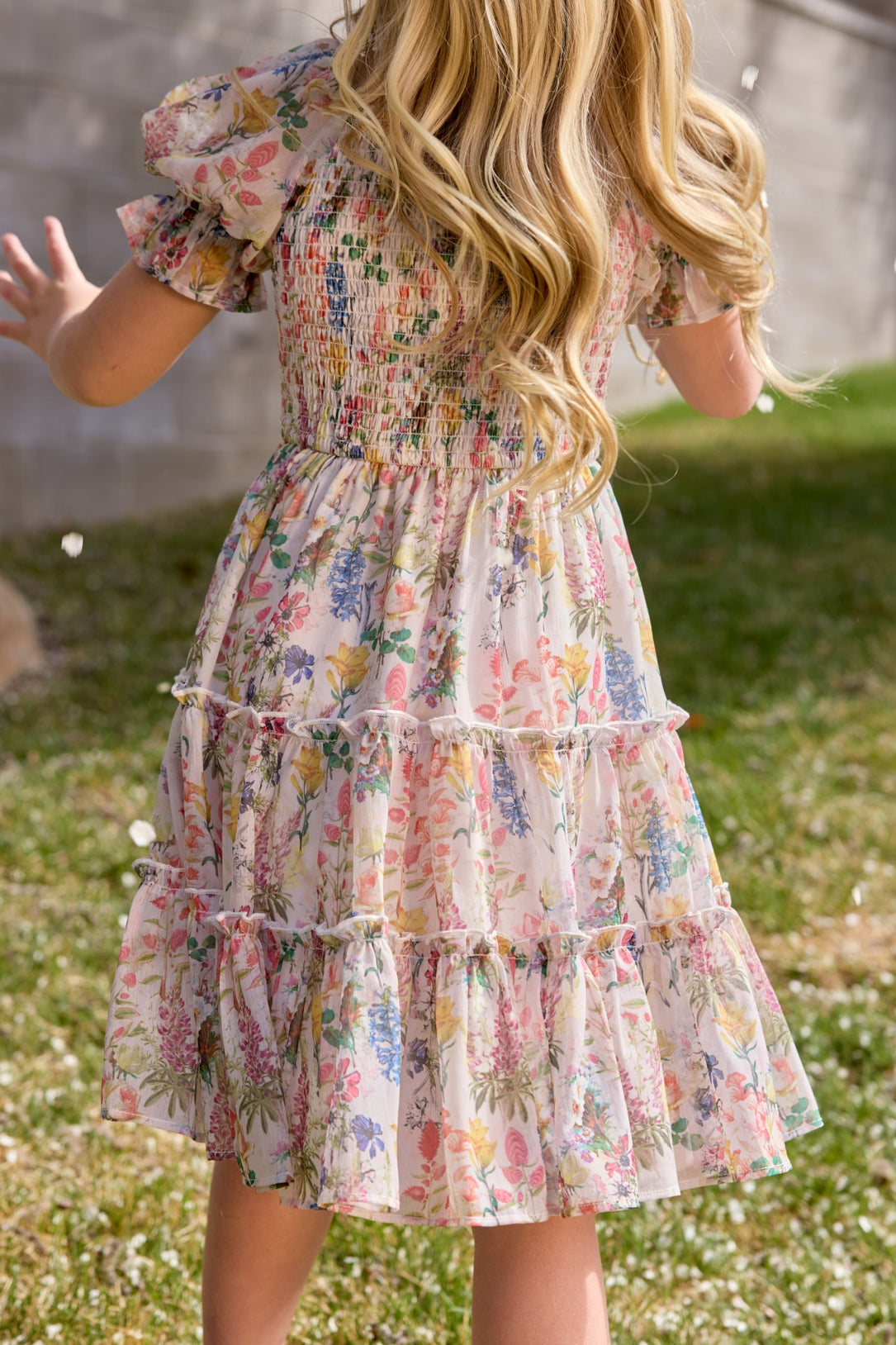 A young girl with long blonde hair wears the Ivy City Co Mini Madeline Dress in Cordelia Floral, standing on grass outdoors, facing away from the camera with her arms slightly outstretched.