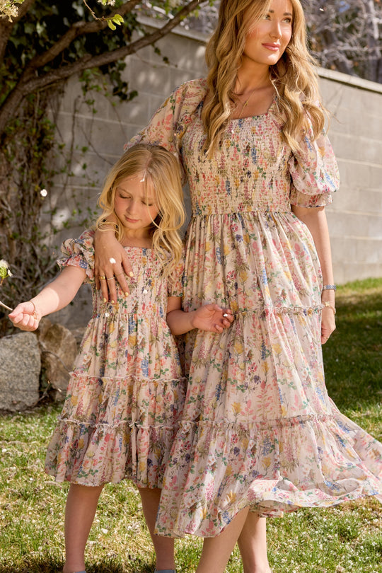 A woman and a young girl stand on grass, holding hands in matching Ivy City Co Cordelia Floral dresses. The girl's Mini Madeline Dress in Cordelia Floral is perfect for a calm, sunny birthday moment together.