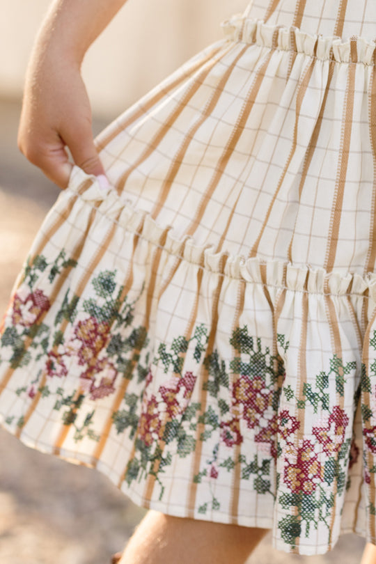 A child’s hand holds the skirt of the Ivy City Co Mini Madeline Dress in Embroidered Cider Grid, which features a charming cream base with tan stripes and an embroidered floral pattern near the hem, adorned with pink and red flowers. The dress is finished with a delicate smocked bodice for added charm.