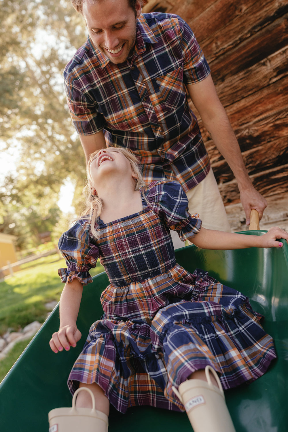 A man pushes a laughing young girl in a green wheelbarrow. Both wear matching plaid outfits, with the girl dressed in Ivy City Co’s Mini Madeline Dress in Midnight Plaid, featuring puff sleeves and a smocked bodice. The scene is outdoors near a wooden building with sunlight filtering through trees.