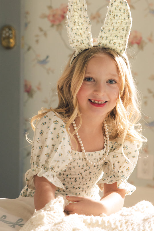 A young girl with blonde hair wears the Ivy City Co Mini Madeline Dress in Blue, paired with a beaded necklace and bunny ear headband. She smiles brightly in warm light against floral wallpaper.