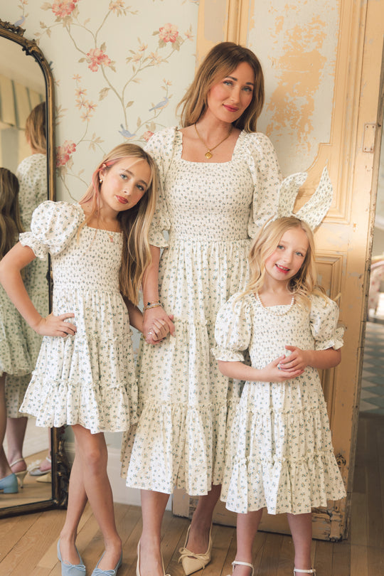 A woman and two girls stand indoors, smiling and holding hands in matching Ivy City Co Mini Madeline Dresses in Blue, featuring small floral prints and puffed sleeves. A vintage mirror and floral wallpaper decorate the background.