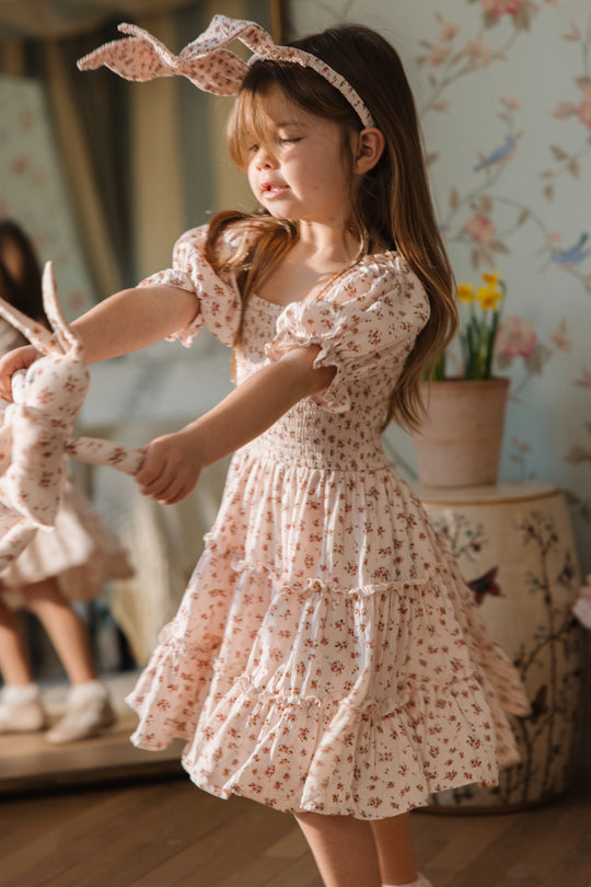 A young girl in an Ivy City Co Mini Madeline Dress in Pink, a light pink floral cotton dress with matching headband, twirls indoors with a fabric bunny amid natural light, floral decor, a mirror, and potted flowers.