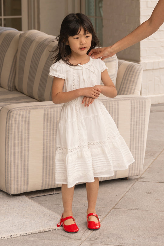 A young girl wears the Ivy City Co Mini Palermo Dress in White with puff sleeves and red shoes, standing indoors near a striped couch as an adult hand reaches toward her face.