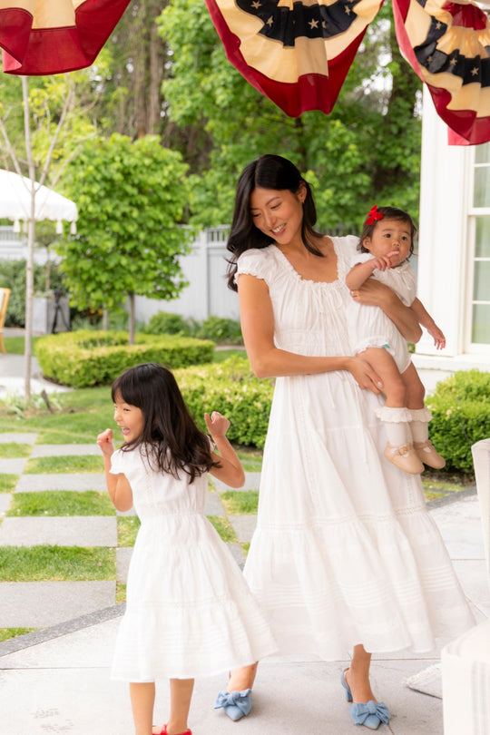A woman in the Ivy City Co Mini Palermo Dress in White smiles while holding a baby and watching a girl twirl. They are outdoors on a patio decorated with greenery and red, white, and blue patriotic bunting overhead.