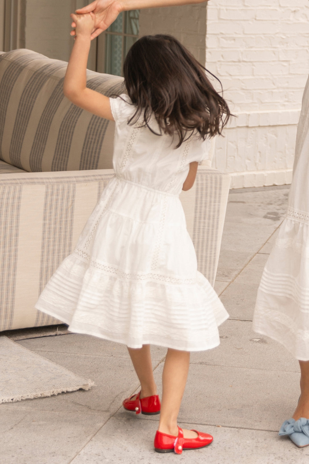 A young girl twirls in the Ivy City Co Mini Palermo Dress in White, holding an adult’s hand, with a striped sofa and white brick wall behind her.