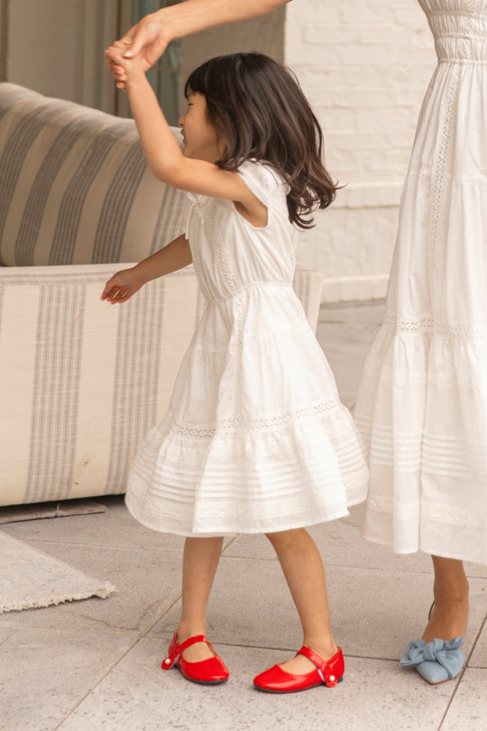 A young girl wears the Ivy City Co Mini Palermo Dress in White as she dances hand-in-hand with an adult, both in white dresses, near a striped sofa.