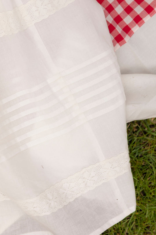 The Mini Palermo Dress in White by Ivy City Co, a cotton dress with horizontal stripes and lace details, is shown laying on grass with a red and white checkered pattern partly visible in the upper right corner.