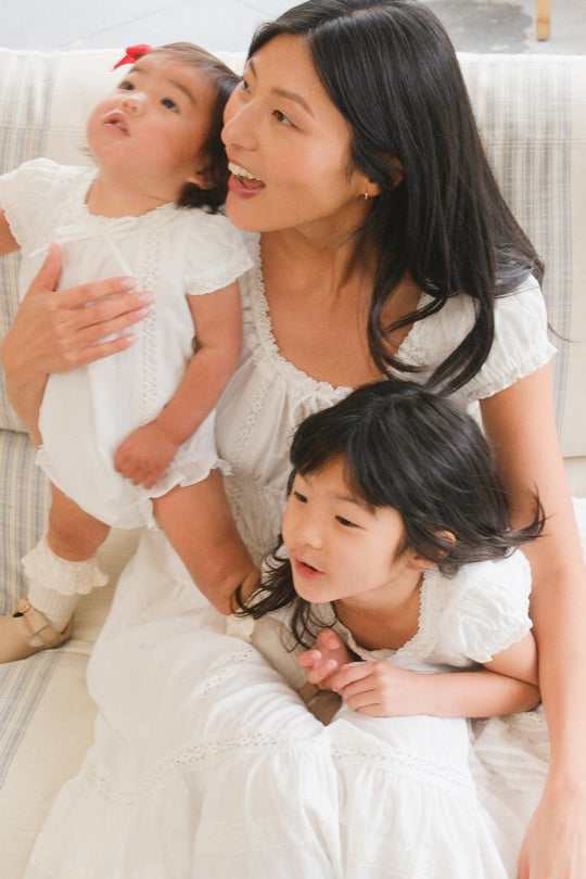 A woman wears the Ivy City Co Mini Palermo Dress in White as she sits on a light couch with two smiling children in white outfits, holding the youngest on her lap while they all look in different directions.