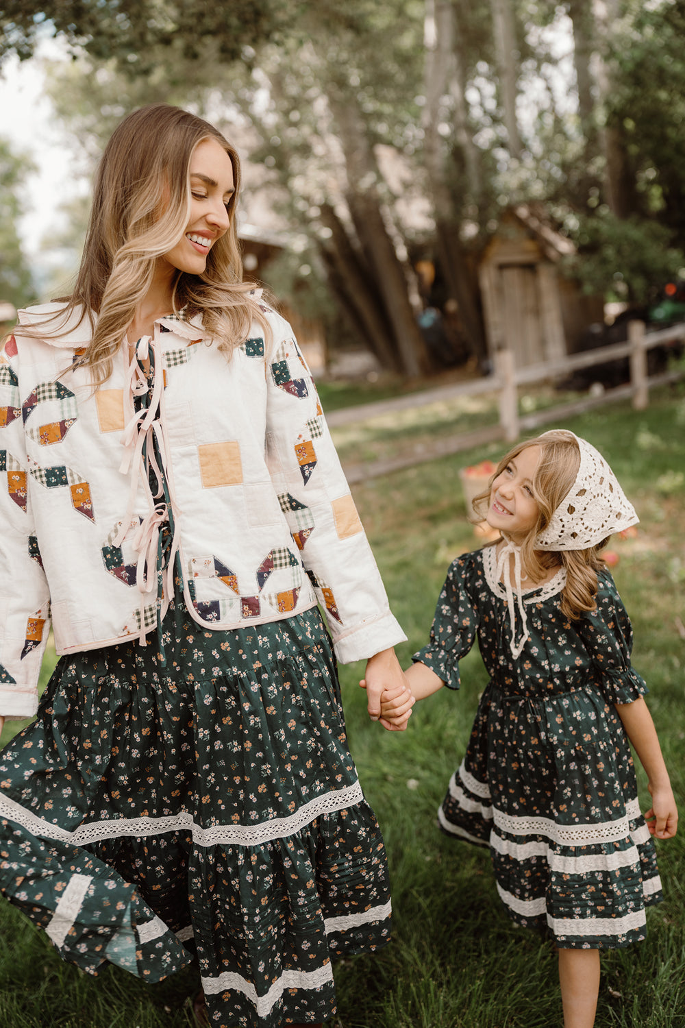 A woman and a young girl, both wearing matching Ivy City Co Mini Palermo 3/4 Sleeves Dresses in Evergreen Lovelorn Ditsy, hold hands and smile at each other while standing on green grass with trees and a wooden fence in the background.
