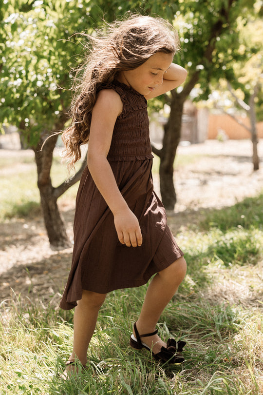 A young girl with long wavy hair walks through grass in a sunlit park, wearing the Ivy City Co Teen Simone Dress in Warm Cocoa. The dress features a smocked bodice and handy pockets, paired with sandals, with trees and soft sunlight in the background.