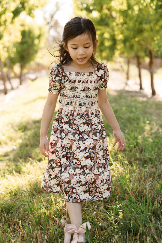 A young girl wearing the Ivy City Co Mini Sunday Best Dress in Midnight Garden, featuring a smocked bodice and puff sleeves, walks outdoors on green grass amid trees and sunlight, looking down with a gentle expression.