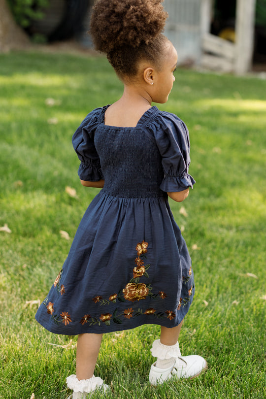 A young girl with curly hair, wearing the Ivy City Co Mini Teagan Dress in Navy Embroidered Fall Floral—which features a smocked bodice and floral embroidery—along with white shoes, stands on green grass in a garden.