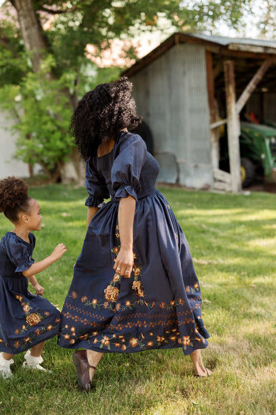 A woman and a young girl, both wearing matching blue cotton dresses with smocked bodices and floral embroidery—such as the Ivy City Co Mini Teagan Dress in Navy Embroidered Fall Floral—hold hands and walk on grass near a rustic shed, surrounded by trees.