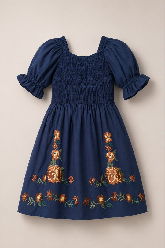The Mini Teagan Dress in Navy Embroidered Fall Floral by Ivy City Co features a classic navy blue cotton design with puffed sleeves, a smocked bodice, and vibrant orange floral embroidery accenting the hem and center for a playful yet timeless look.