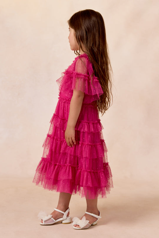 A young girl with long brown hair, wearing the Ivy City Co Mini Whimsical Dress in Fuchsia and white sandals, stands sideways against a light beige background.