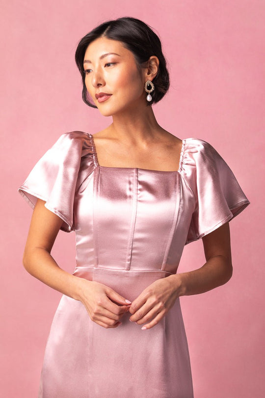 A woman wearing the Maddie Dress in Blush Satin by Ivy City Co, featuring flutter sleeves and a lustrous finish, styled with pearl earrings and dark hair against a pink background—ideal for bridesmaids or as an elegant maxi event dress.