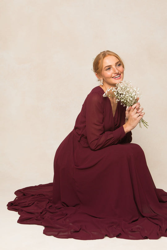 A woman wearing the Ivy City Co Andie Dress in Wine Chiffon kneels on the floor, holding a small bouquet of white flowers and smiling softly against a plain light background.