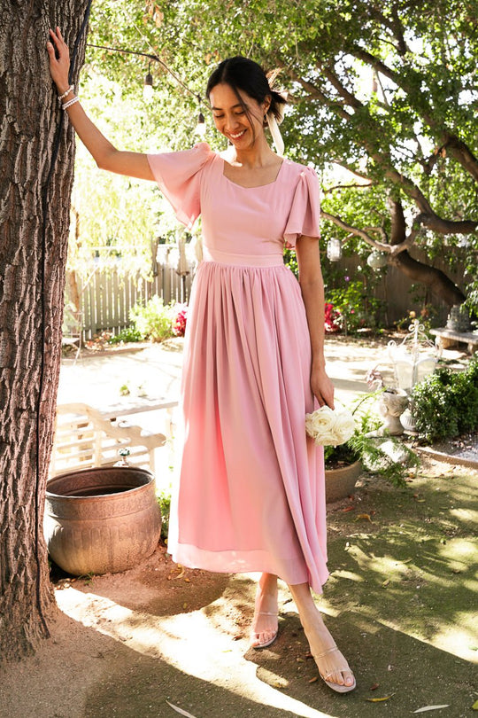 A woman wearing the Abbie Dress in Blush by Ivy City Co stands by a tree in a sunlit garden, holding white flowers and smiling, with greenery and a bench visible in the background.