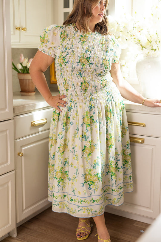 A woman stands in a sunlit kitchen, one hand resting on the counter, wearing the Jennifer Dress in Garden Tea Party Floral by Ivy City Co—a lightweight, floral print design—paired with yellow strappy sandals as sunshine pours in through the window behind her.