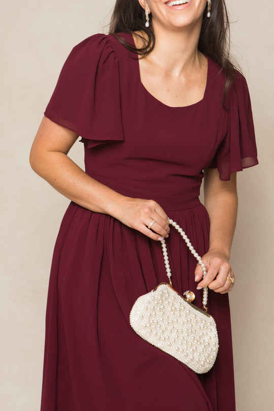 A woman smiles while wearing the Ivy City Co Abbie Dress in Wine Chiffon and holding a white beaded handbag. Her dark hair is down, and she accessorizes with pearl earrings against a neutral background.