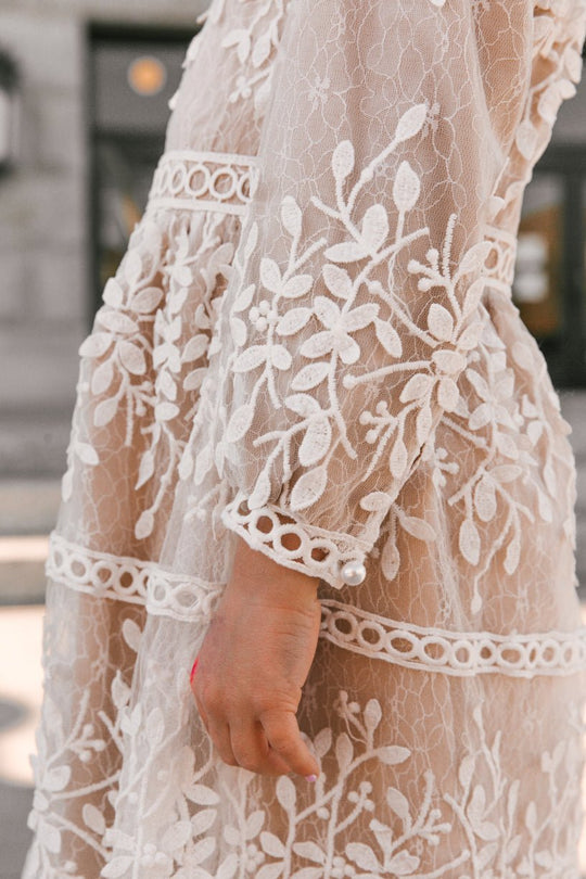 A close-up of a person wearing the Ivy City Co Mini Sicily Dress with Long Sleeves—a sheer, champagne-lined white lace dress featuring floral embroidery and circular trim. The person’s hand, with pink nail polish, is visible in the foreground while the background is blurred.