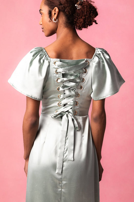 An elegant woman models the Maddie Dress in Sage Satin by Ivy City Co, featuring puff sleeves and a striking lace-up corset back. She faces away against a pink backdrop, highlighting the dress’s shiny, light green sage satin fabric.