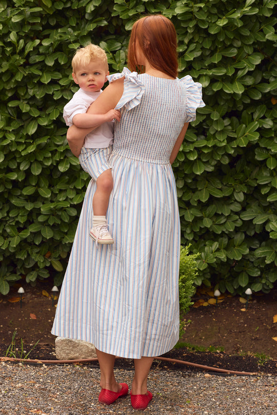 A woman with long red hair, wearing the Ivy City Co Hattie Dress in Heritage Stripe, stands outdoors with her back to the camera, holding a young blond child in matching stripes by green leafy bushes.