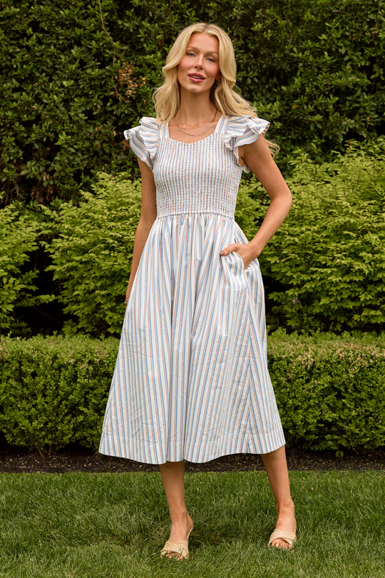 A woman with long blonde hair stands on grass in front of green bushes, wearing the Ivy City Co Hattie Dress in Heritage Stripe—a nursing-friendly midi dress with blue and tan vertical stripes, ruffled sleeves, pockets, and beige sandals.