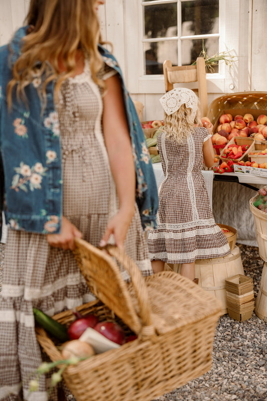 Two women wearing Ivy City Co’s Mini Palermo Dress in Hickory Gingham shop at an outdoor fruit stand. One holds a wicker basket filled with vegetables, while the other, paired with a bonnet and reaching for apples, stands beside her. Sunlight streams through a window and wooden crates fill the background.