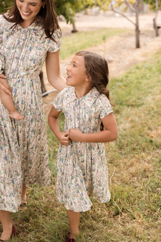 A smiling woman and a young girl, both wearing matching floral dresses, walk together outdoors on a grassy path. The woman wears a viscose midi dress while the young girl is dressed in the charming Mini Amber Dress in Autumn Bouquet by Ivy City Co. The girl looks up at the woman with admiration, and trees are visible in the background.