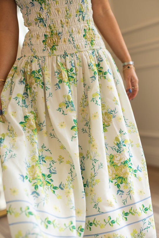A person wearing the Jennifer Dress in Garden Tea Party Floral by Ivy City Co, a maternity-friendly white dress adorned with yellow and green floral patterns, featuring a smocked bodice and flowing skirt crafted from lightweight fabric. The photo shows the dress from the waist down.