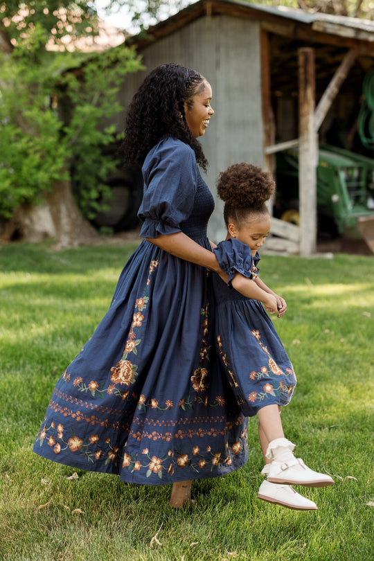 A woman and a young girl, both wearing matching Ivy City Co Mini Teagan Dresses in Navy Embroidered Fall Floral with charming balloon sleeves and a smocked bodice, hold hands and smile as they walk barefoot on green grass in front of a rustic shed.