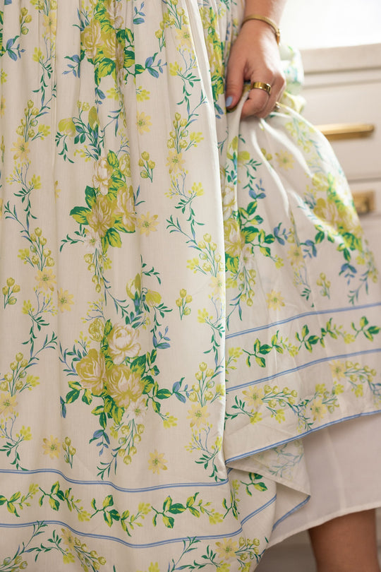 A close-up of a person wearing the Jennifer Dress in Garden Tea Party Floral by Ivy City Co, featuring a white base with yellow and green floral patterns, standing indoors. The lightweight fabric drapes softly as the person’s hand, adorned with gold rings, gently holds the skirt.