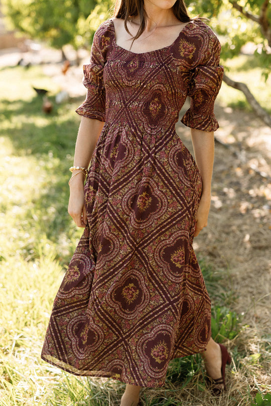 Wearing the Ivy City Co Josephine Dress in Nocturne Diamond Rose, a woman stands on grass outdoors with her face partially out of frame. Sunlight highlights the patterned maroon and gold cotton fabric and garden surroundings, drawing attention to the dress’s smocked bodice and maternity-friendly silhouette.