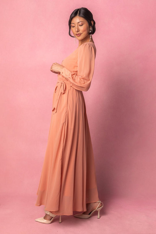 A woman wearing the Ivy City Co Andie Dress in Apricot Crush, styled with pearl earrings and beige heels, stands against a pink background, looking to the side.