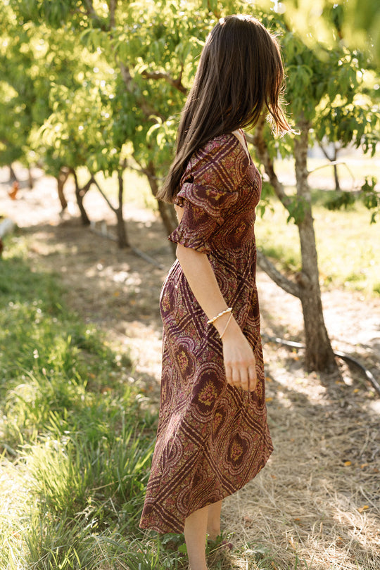 A woman stands barefoot outdoors among trees, her head turned to the side and hair partially covering her face. Sunlight filters through the leaves, highlighting the smocked bodice of her maternity-friendly Josephine Dress in Nocturne Diamond Rose by Ivy City Co.