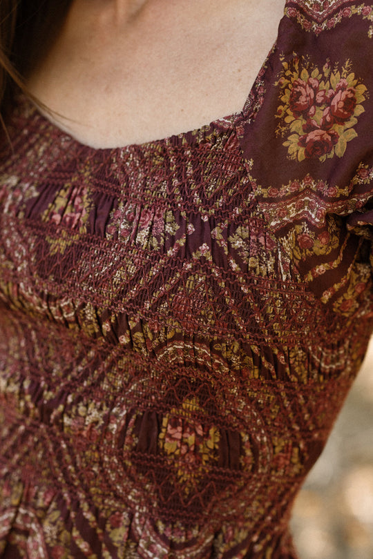 Close-up of a person wearing the Ivy City Co Josephine Dress in Nocturne Diamond Rose—a burgundy cotton dress featuring a diamond rose floral pattern, intricate stitching, and a smocked bodice. Only the upper torso, neckline, and shoulder are visible, highlighting its maternity-friendly design.