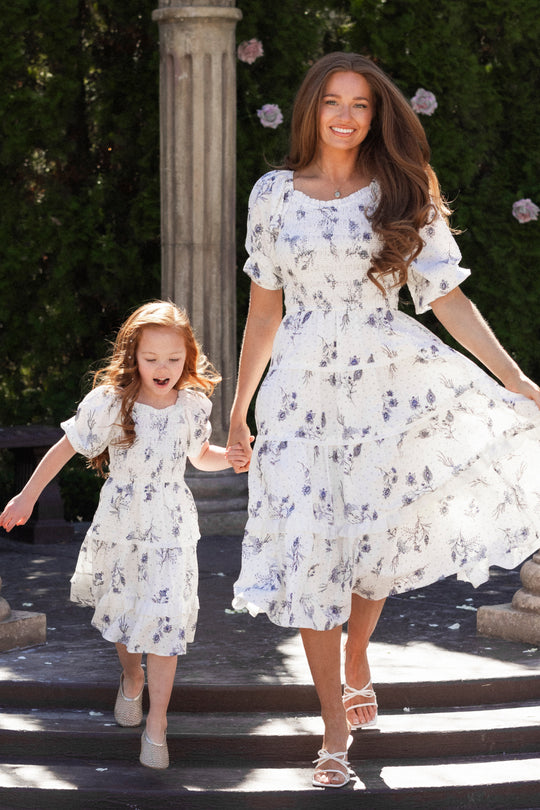 A woman and a young girl wear matching Ivy City Co Roselyn Dresses in Blue Enchanted Floral, holding hands and smiling as they walk outdoors near pillars and greenery. The woman's dress is maternity-friendly and flows in the breeze.