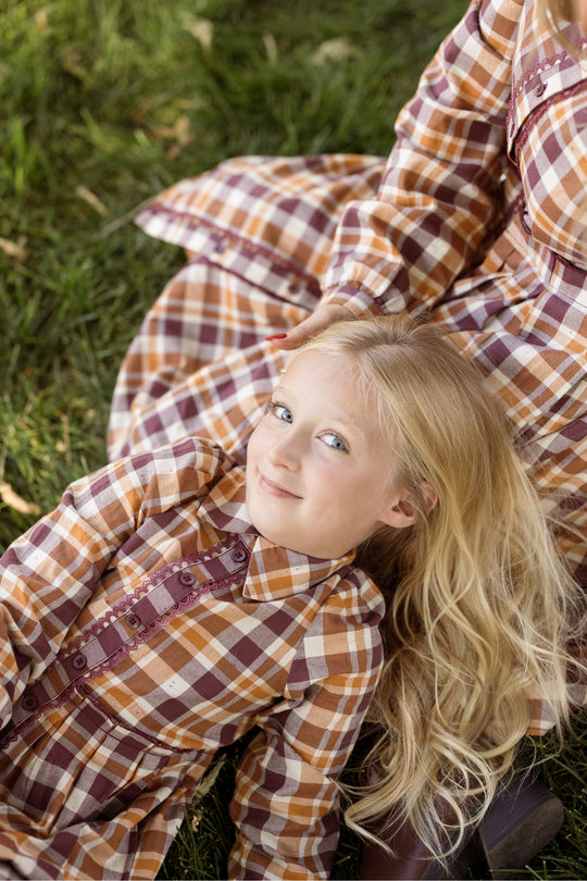 A young girl with long blond hair, wearing the Ivy City Co Mini Bowie Long Sleeve Dress in Spiced Plaid—featuring charming balloon sleeves in brown and orange hues—lies smiling on green grass, looking up at the camera. Beside her, another person sits in a matching dress.