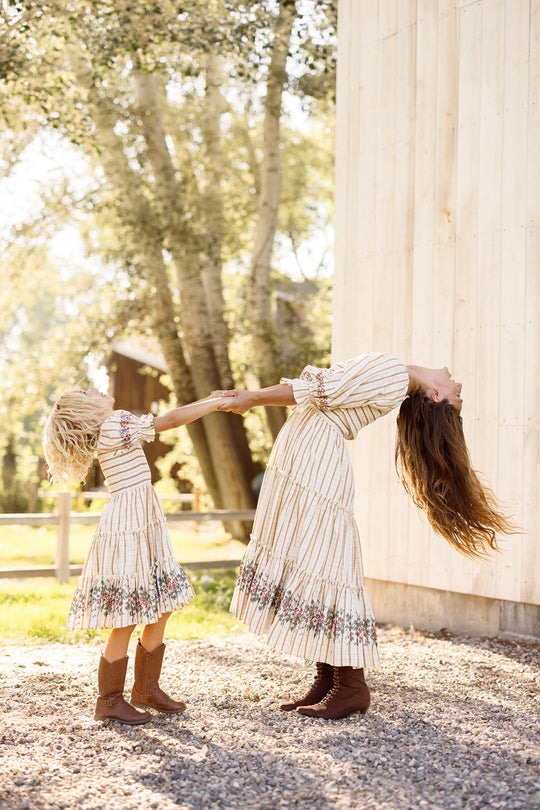 An adult and a young girl, both dressed in Ivy City Co’s Mini Madeline Dress in Embroidered Cider Grid featuring smocked bodices and playful puff sleeves, hold hands and lean back, laughing together outdoors near a wooden wall and trees on a sunny day.