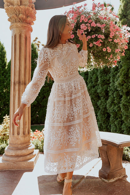 A woman wearing the Ivy City Co Sicily Dress in Champagne Lining stands outside by a stone column, smiling as she leans toward a large basket of pink flowers on a sunny day.