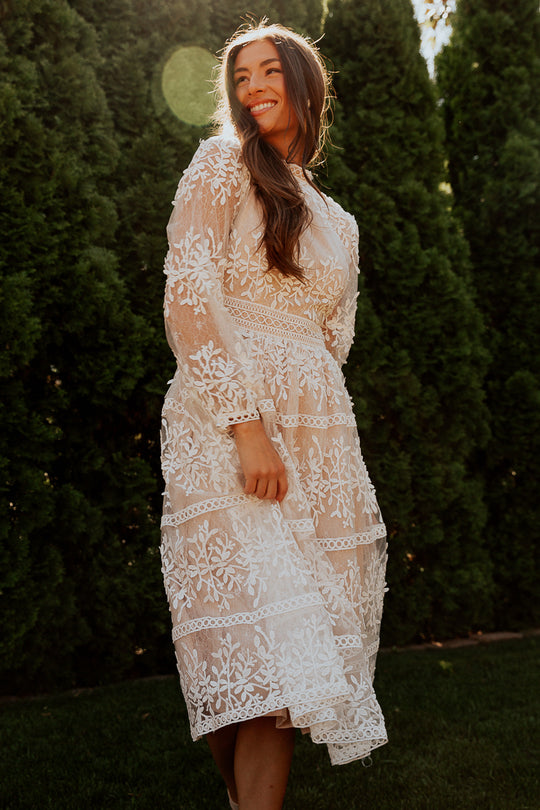 A woman in the Ivy City Co Sicily Dress in Champagne Lining, with a white floral overlay and long sleeves, stands outside smiling by tall green trees as sunlight filters through the scene.