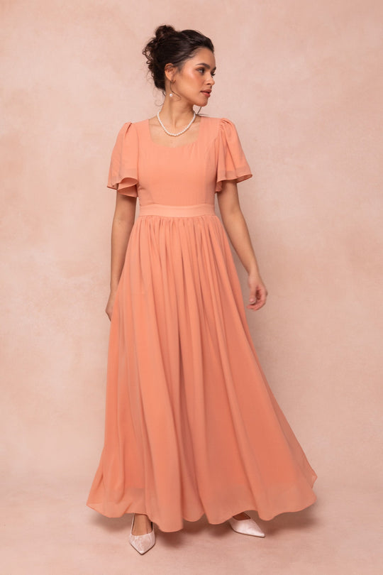 A woman wearing the Ivy City Co Abbie Dress in Apricot Crush, a short-sleeved maxi dress, stands against a soft beige background looking to her left. She accessorizes with pearl earrings, a necklace, and pointed-toe shoes.