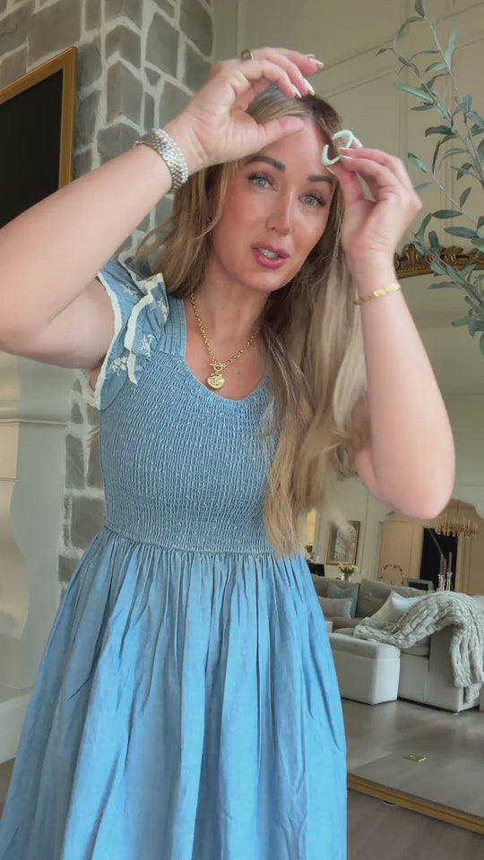 A woman stands indoors in a stylish living room with neutral decor, smiling softly as she holds a small white ribbon above her head. She wears the Hattie Ankle Length Dress in Embroidered Chambray by Ivy City Co—a maternity-friendly piece that effortlessly blends comfort and elegance with its blue smocked bodice design.